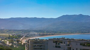 Paisagem costeira com montanhas ao fundo, uma praia e um edifício moderno com vários andares.