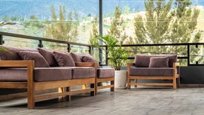 Un balcón con un sofá y una silla de madera, una planta en maceta y vistas a árboles y montañas.