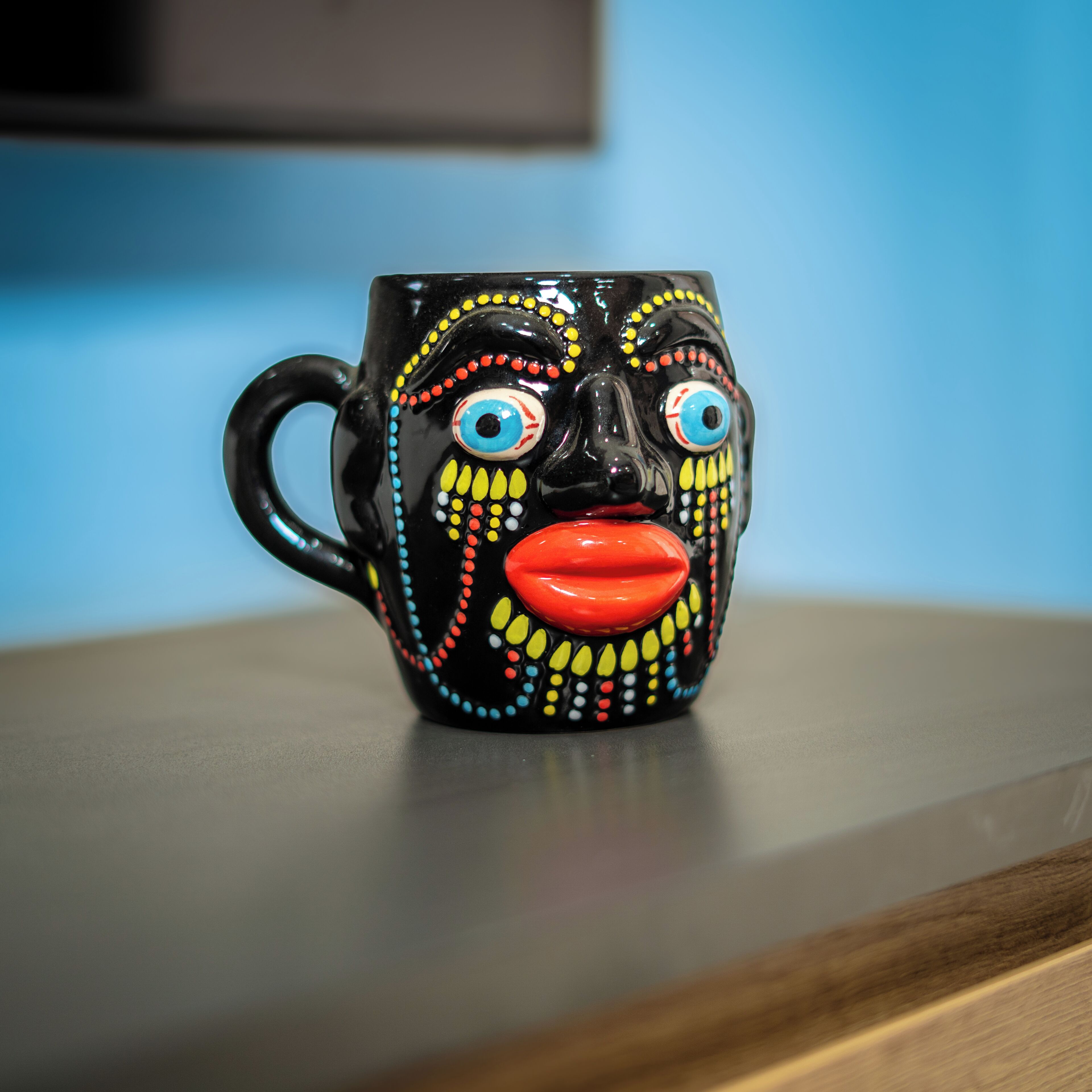 Una tazza nera con un motivo colorato a forma di viso su un piano di legno.