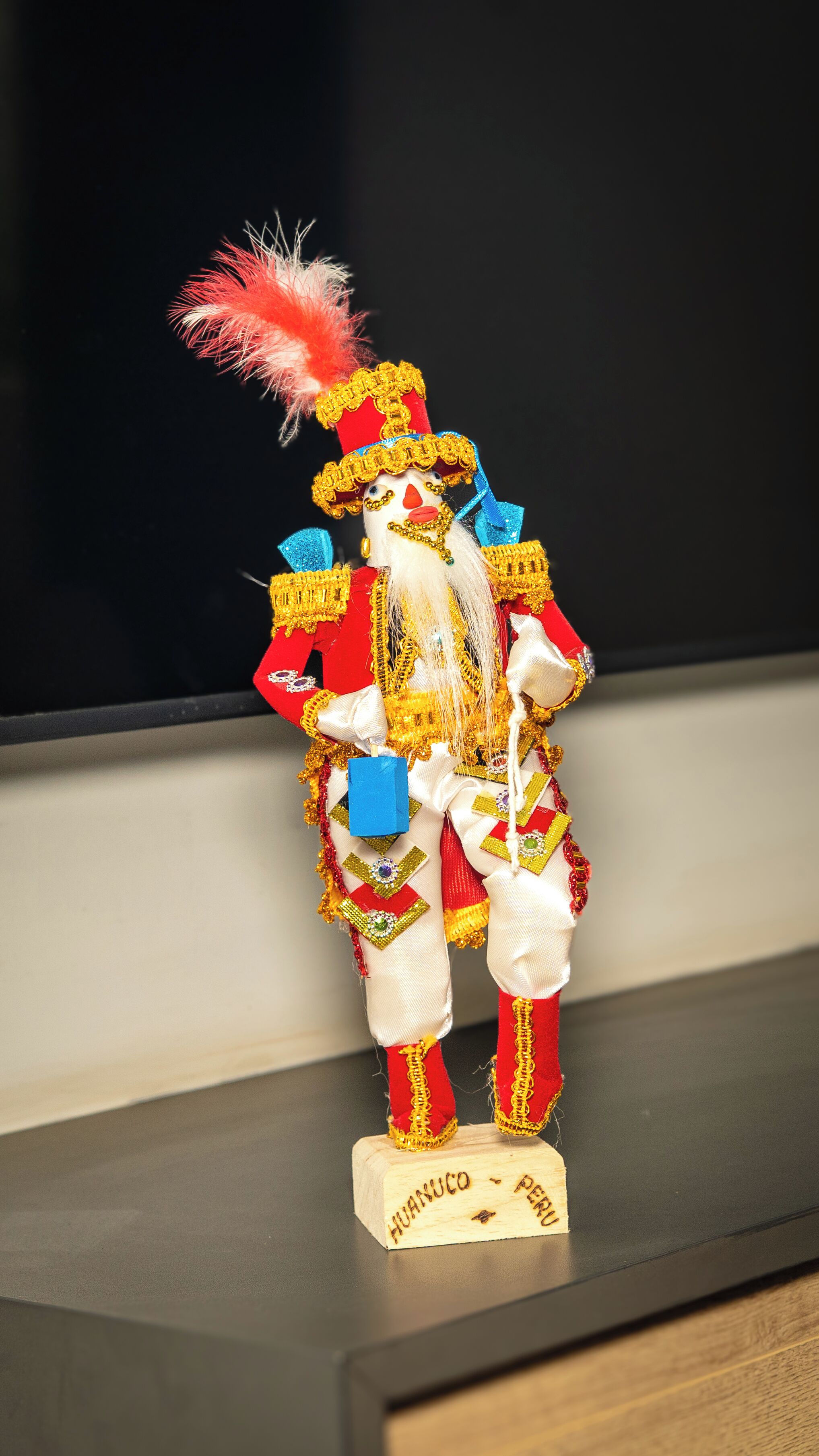 Una figurita de madera de una persona con sombrero rojo y una cinta azul, sobre un base de madera con la leyenda 'HUANUCO PERU'.