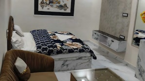 Un dormitorio moderno con cama, sofá, mesa de centro de cristal, ventilador de techo, iluminación empotrada, televisor de pared y una obra de arte enmarcada.