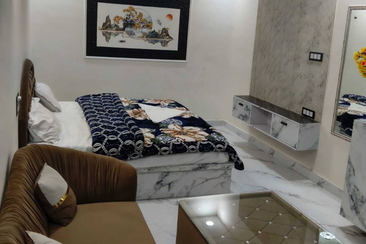 A modern bedroom with a bed, a sofa, a glass coffee table, a ceiling fan, recessed lighting, a wall-mounted TV, and a framed artwork.