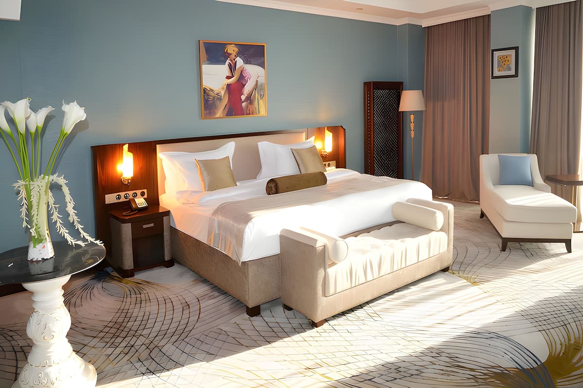 Habitación de hotel con una cama grande, mesitas de noche, una silla, un florero con flores y un cuadro en la pared.