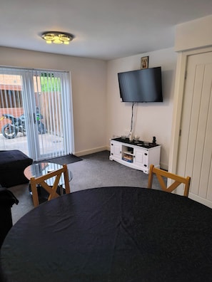 A room with a round table, two wooden chairs, a television mounted on the wall, a sliding glass door leading outside, and a white cabinet with a television on top.