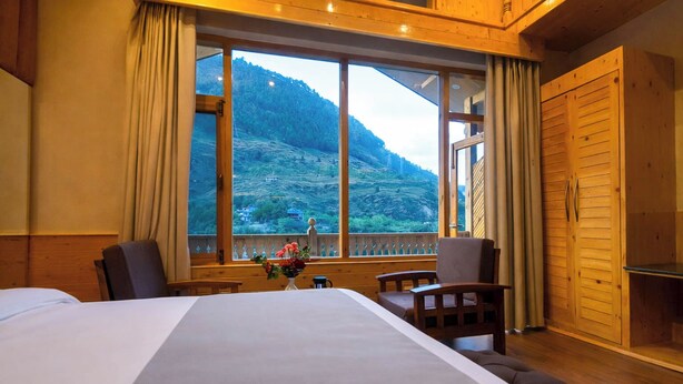 Ein Hotelzimmer mit einem großen Bett, einem Holztisch, einem Stuhl und einem Fenster mit Blick auf die Berge.