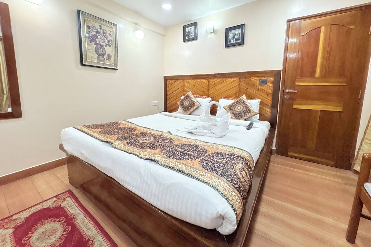 Una habitación de hotel con cabecera de madera, una cama grande con colcha estampada, una alfombra roja y beige, y una puerta de madera.