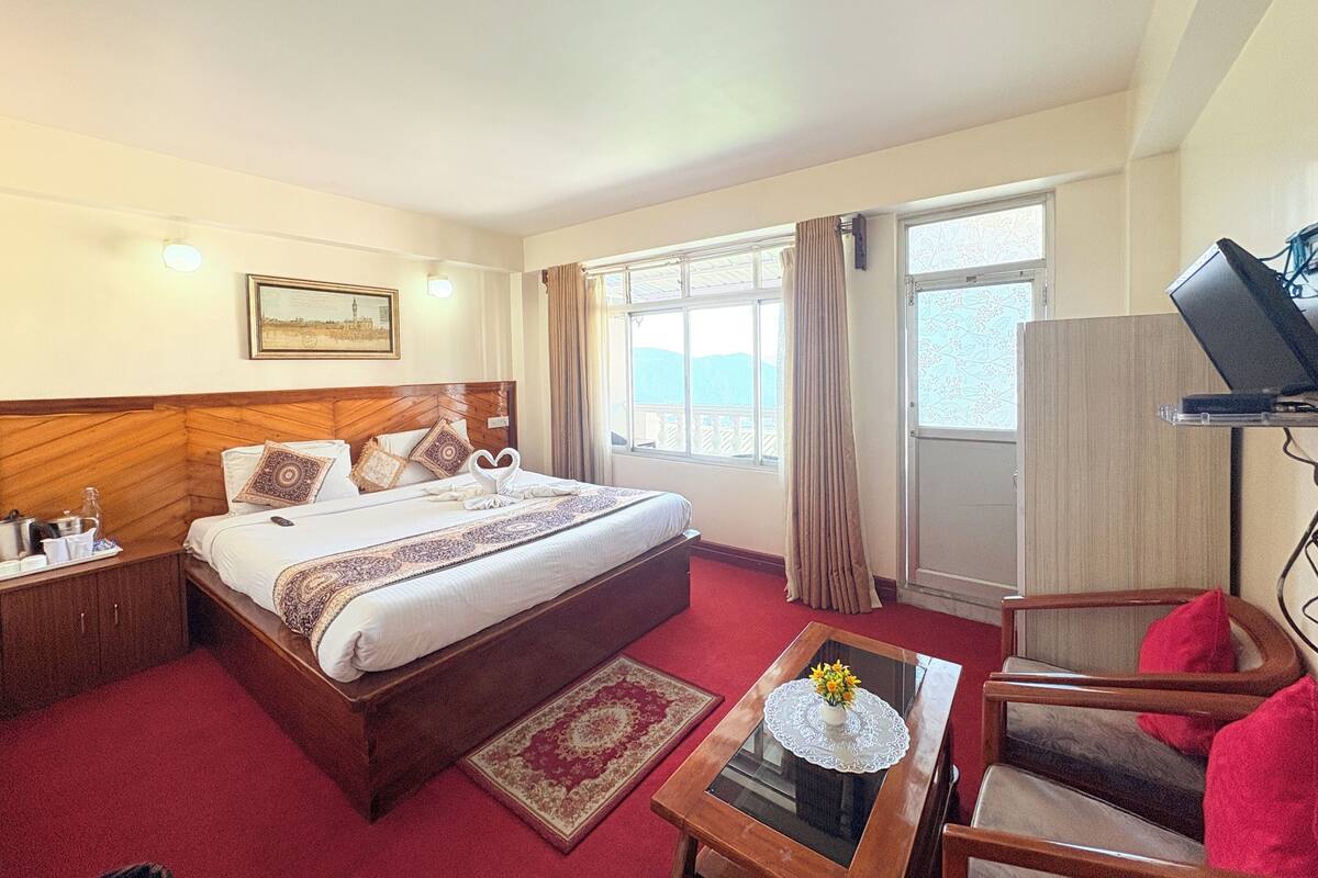 Habitación de hotel con una cama grande, un escritorio con un florero, un televisor sobre un mueble bajo y una ventana con vistas a las montañas.