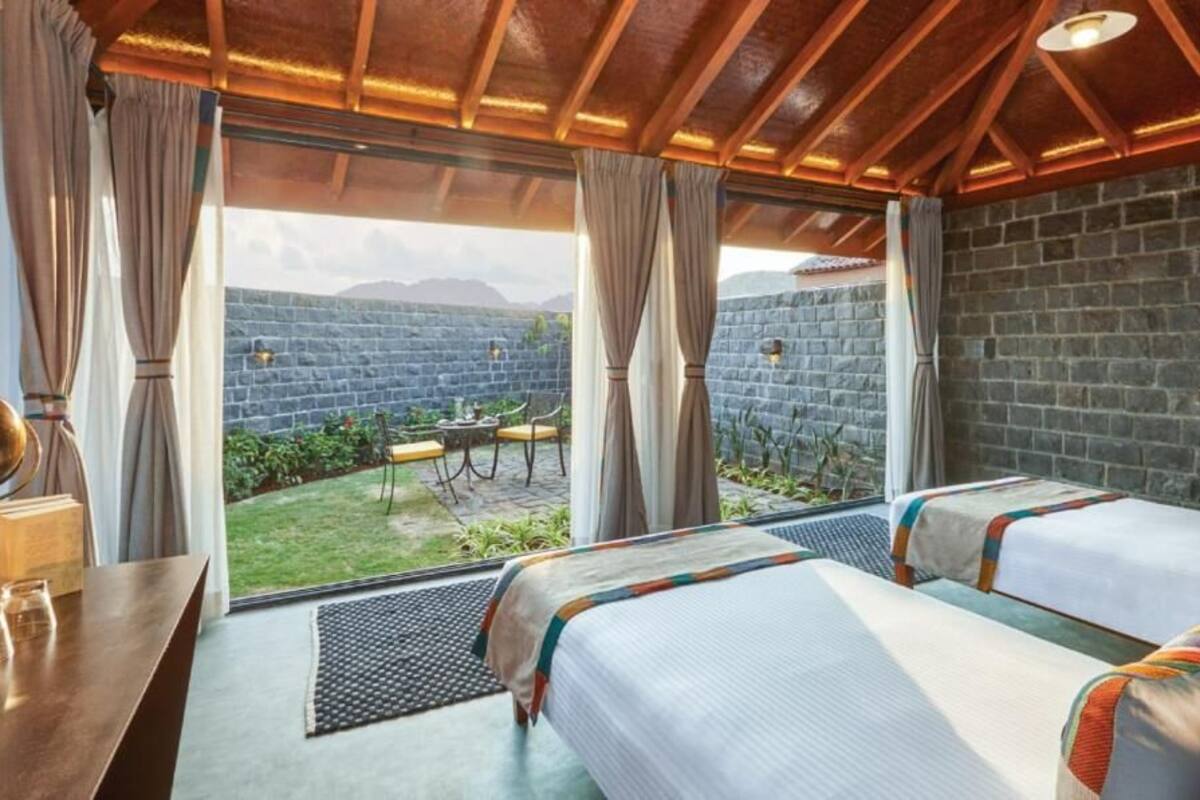 A room with two beds, a table, and a view of a garden and mountains.