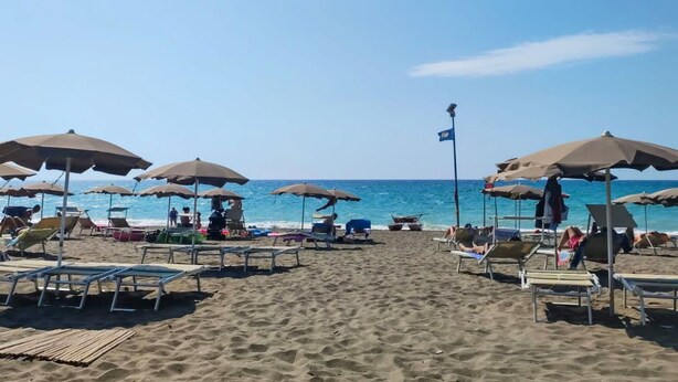 Une plage avec des transats et des parasols, un mât de drapeau, et l’océan en arrière-plan.