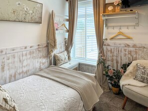 Ein Schlafzimmer mit einem Bett, einem Sessel, einem Fenster mit Vorhängen, einem Regal mit einem Schild und einer Wand mit einem Blumenbild.