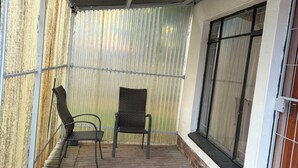 A covered outdoor area with a brick floor, two wicker chairs, a metal railing, and a window with a metal grill. - Perfect 3 x 1-bedroom Rooms in beautiful Sasolburg for your getaway (Sasolburg)