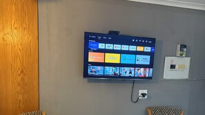 A room with two patterned armchairs, a small wooden table, a TV mounted on the wall displaying various streaming apps, and a power outlet. - Perfect 3 x 1-bedroom Rooms in beautiful Sasolburg for your getaway (Sasolburg)