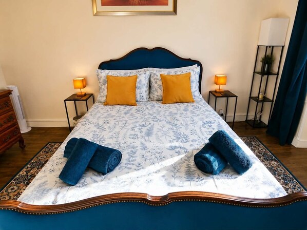 Een bed met een bloemenprint sprei, twee kussens en twee opgerolde handdoeken.