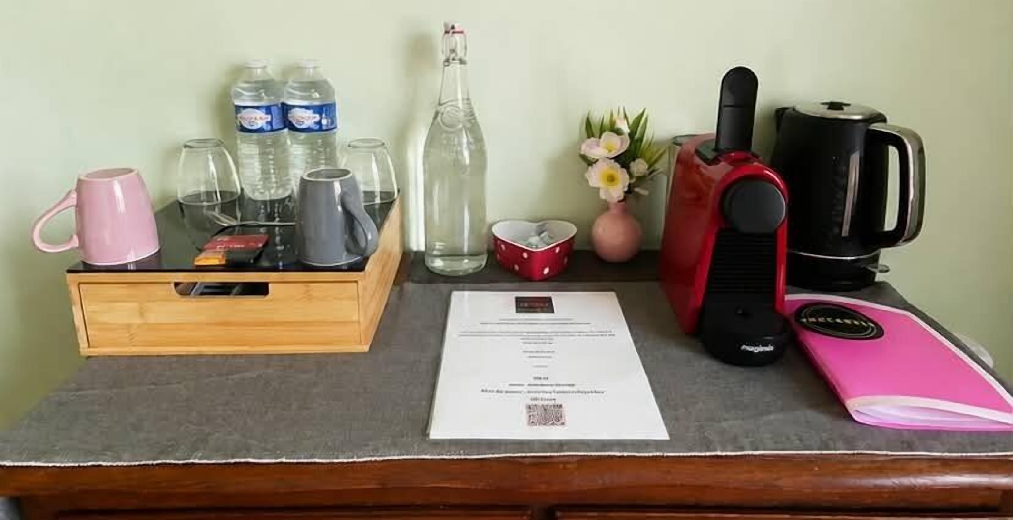 A wooden table with a tray holding mugs, bottles, and glasses, a coffee maker, a pink notebook, and a document.