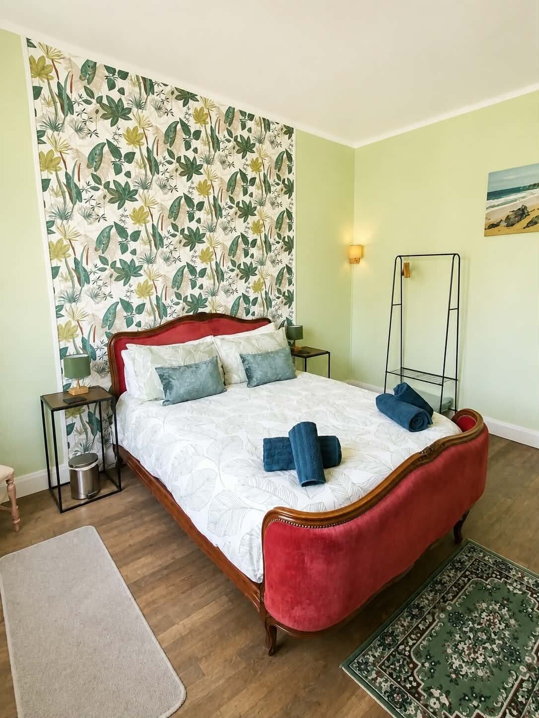 A bedroom with a floral wallpaper, a red upholstered bed, a bedside table with a lamp, and a small rug.