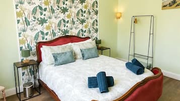 A bedroom with a floral wallpaper, a red upholstered bed, a bedside table with a lamp, and a small rug.