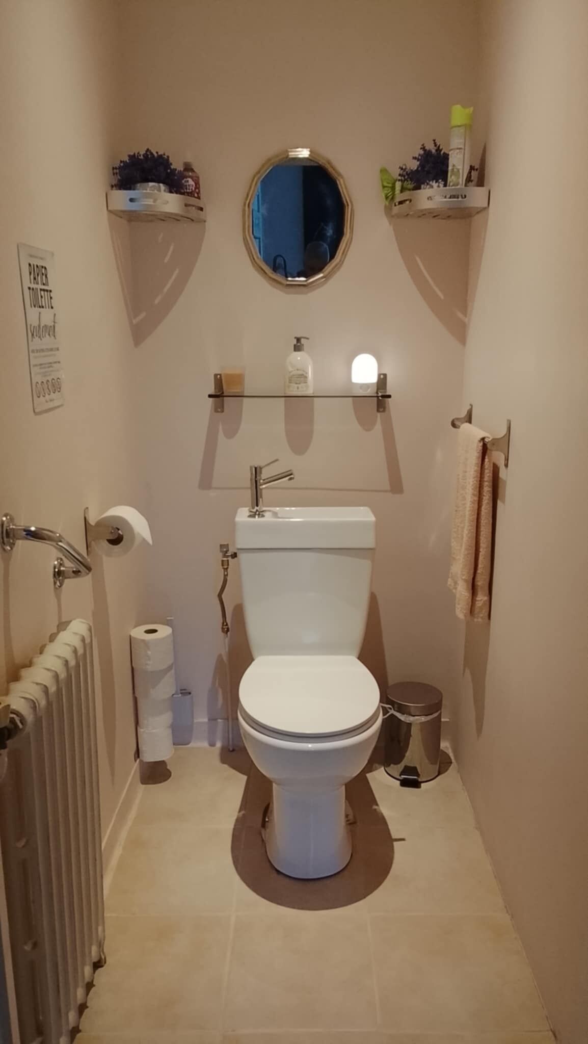 A small bathroom with a toilet, a radiator, a roll of toilet paper, a trash can, and a mirror.