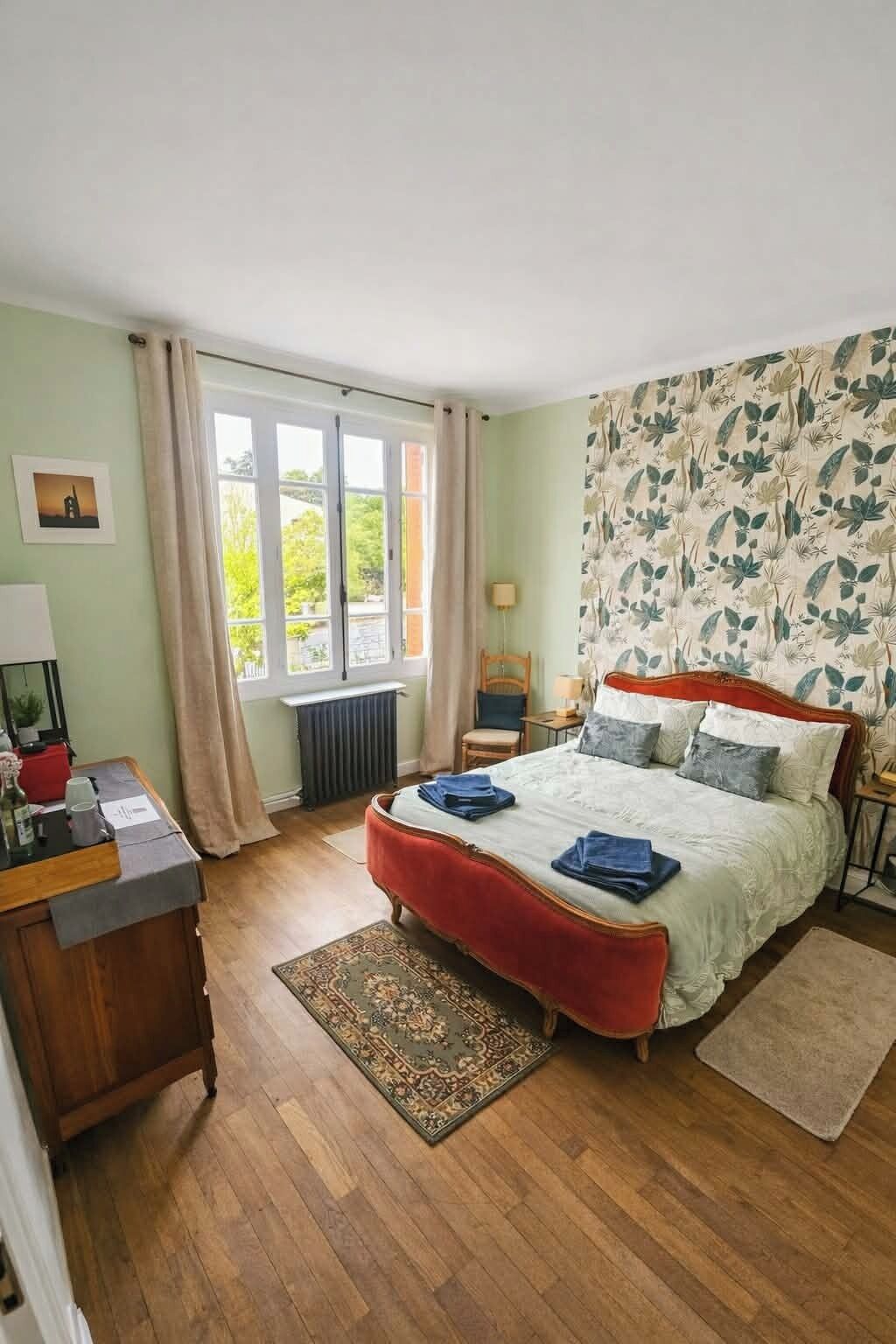 A bedroom with a large bed, a desk, a chair, a radiator, and a window with curtains.