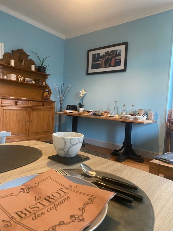 A dining area with a table set for two, a wooden cabinet, a framed picture on the wall, and a small table with glassware and food items. - Trecastle Chambre d'Hotes (Châlus)