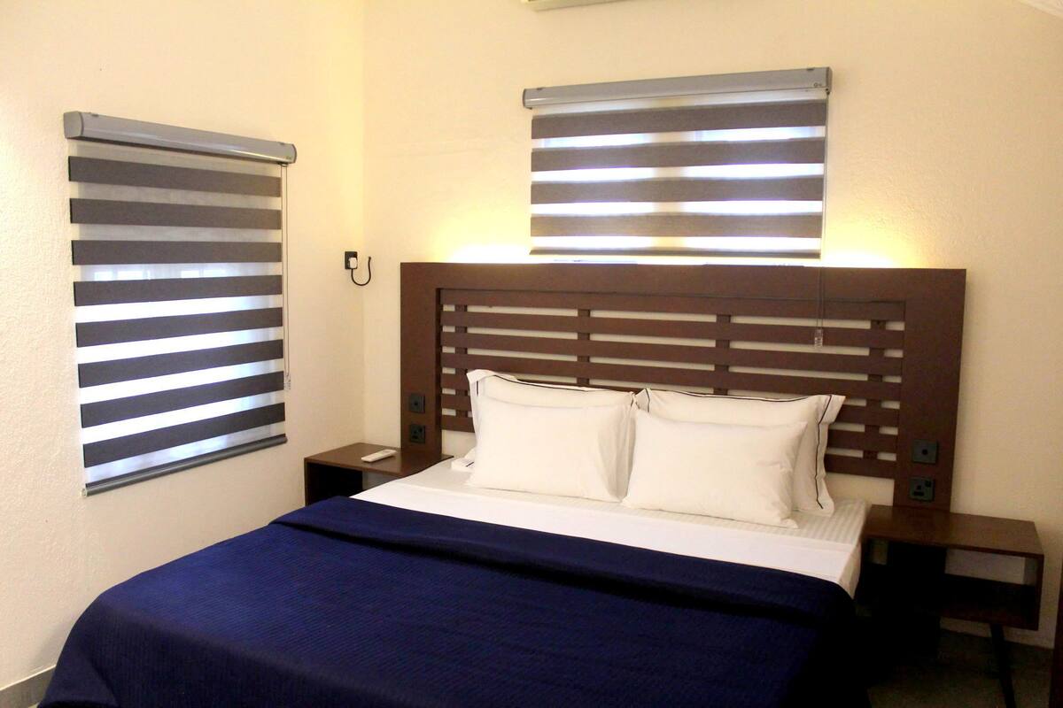 Una habitación de hotel con una cama, dos mesitas de noche y dos ventanas con persianas.