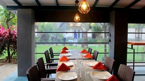 A dining area with a table set for six, surrounded by wicker chairs, with a view of a garden through large windows. - Morning Star Resort Pvt Ltd (Negombo)