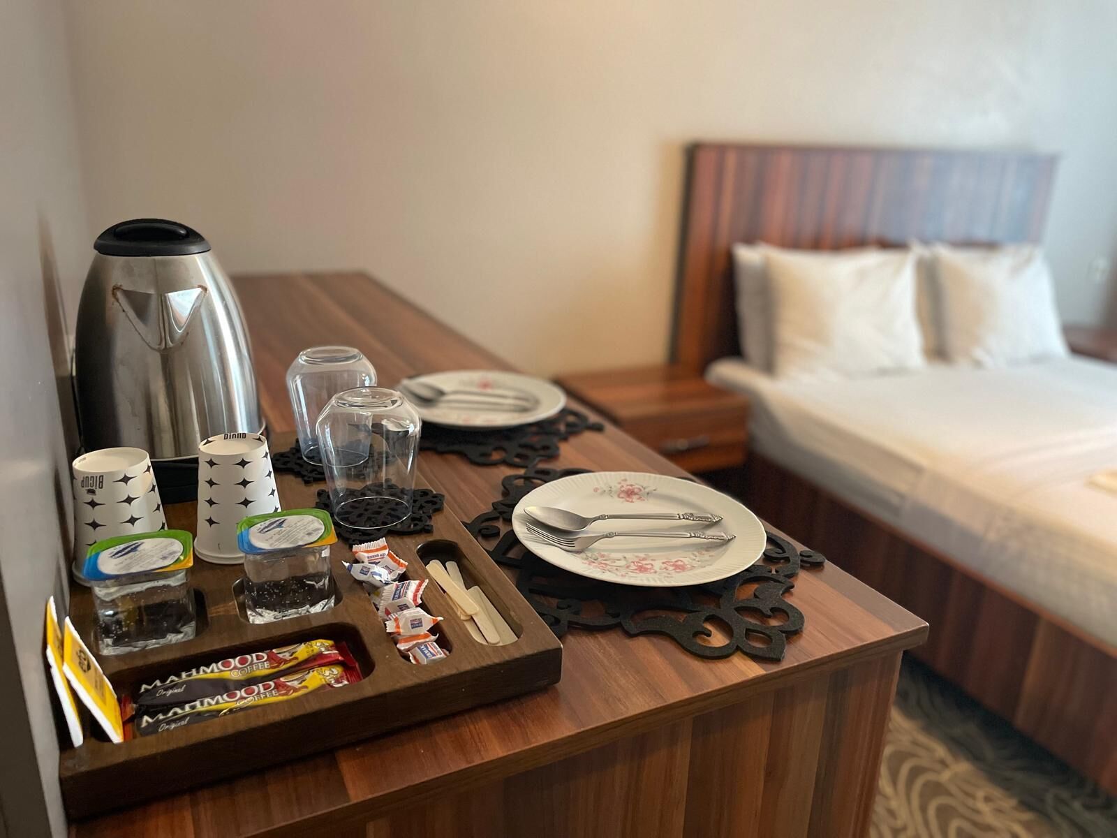 A hotel room with a wooden table set with a coffee maker, cups, glasses, and a tray with coffee packets.