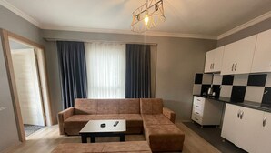 Un espace de vie comprenant un canapé, un pouf et une petite table. On y trouve également une cuisine avec des placards et un plan de travail. Une fenêtre avec des rideaux est visible.