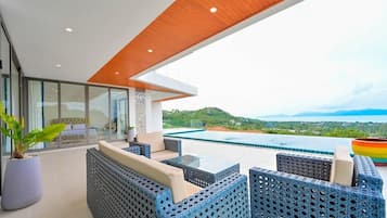 Un área de estar moderna al aire libre con mobiliario de mimbre y una mesa de vidrio, con vistas a una piscina y un paisaje pintoresco.