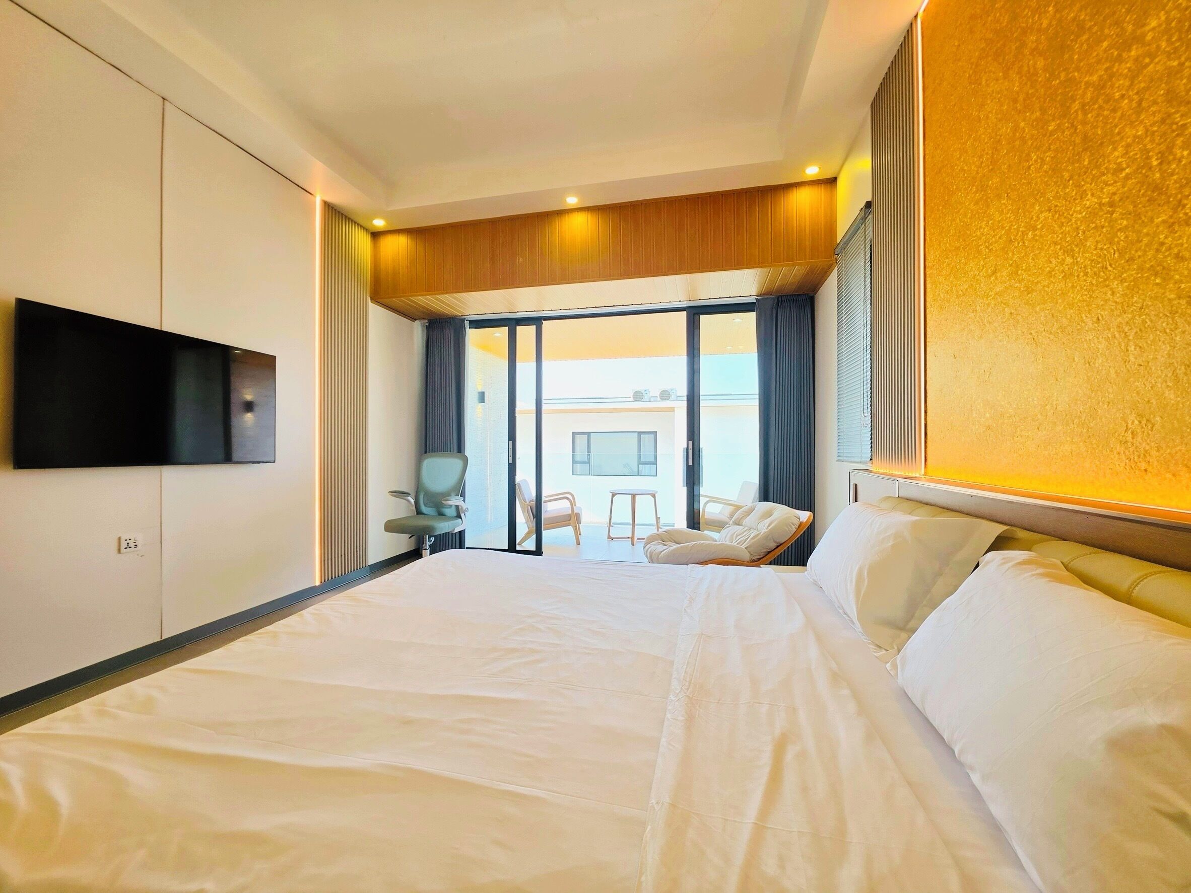 A hotel room with a large bed, a flat-screen TV mounted on the wall, a seating area with chairs, and a view of the outside.