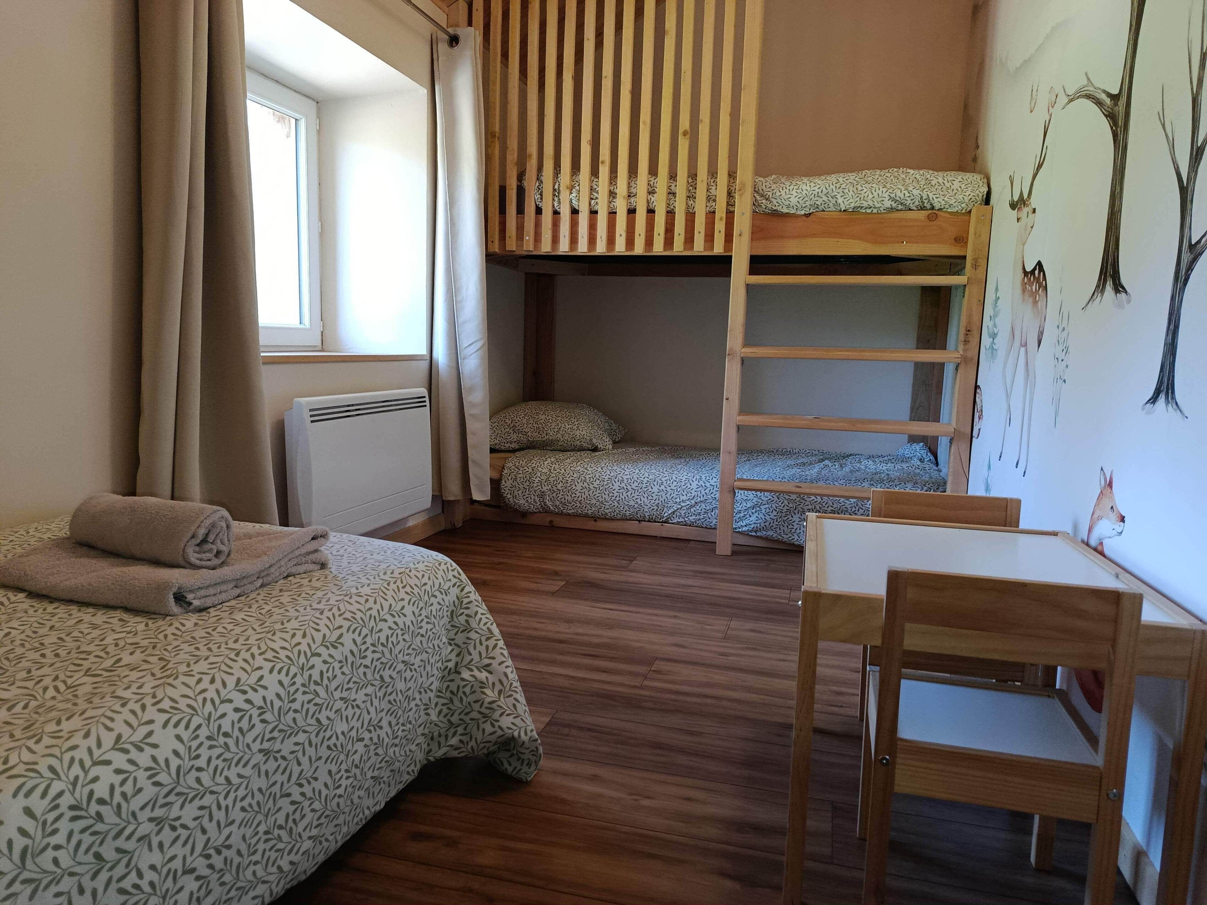 Een kamer met twee bedden, een stapelbed, een tafel, een stoel, een radiator en een raam met gordijnen.