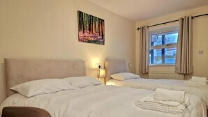 A hotel room with two beds, a headboard, a bedside table, a lamp, a window with curtains, and a painting on the wall.