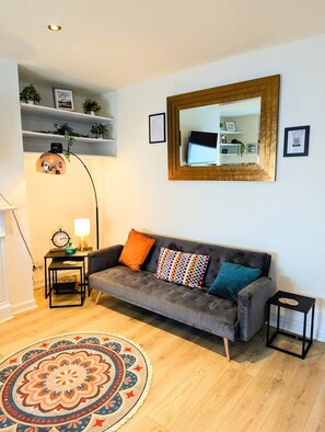 Un salon avec un canapé gris, des coussins colorés, un tapis rond à motifs, un grand lampadaire et un grand miroir accroché au mur.