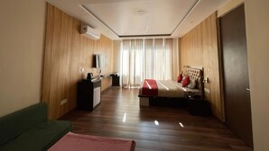 A modern hotel room with a wooden floor, a bed with red and white bedding, a green sofa, a desk with a flat-screen TV, and a large window with curtains. - Indra Stay by Panache Hotels (Baijnath)