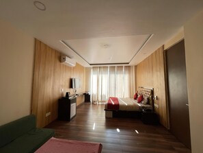 A modern hotel room with a wooden floor, a bed with red and white bedding, a green sofa, a desk with a flat-screen TV, and a large window with curtains.