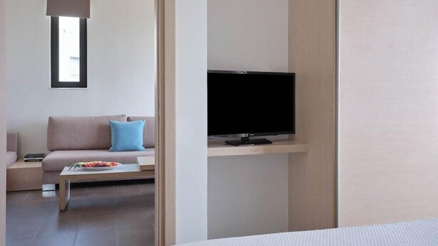 A modern hotel room with a bed, a television on a stand, a sofa with cushions, and a small table with a flower arrangement. - Atlantica Mikri Poli Rhodes (Rhodes)