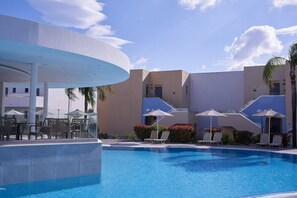 Ein Schwimmbecken mit Liegestühlen und Sonnenschirmen, umgeben von Gebäuden im modernen Architekturstil.
