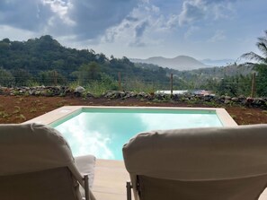 Ein Pool mit Liegestühlen im Vordergrund, umgeben von einem Garten mit Steinen und Pflanzen, mit Blick auf ferne Hügel und Berge.
