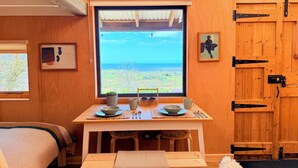 Una habitación acogedora con un ventanal grande, paredes de madera, una cama, una mesita con una taza y un plato, y un cuadro enmarcado.