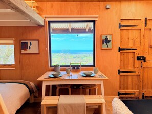 A cozy room with a large window, wooden walls, a bed, a small table with a cup and bowl, and a framed picture.