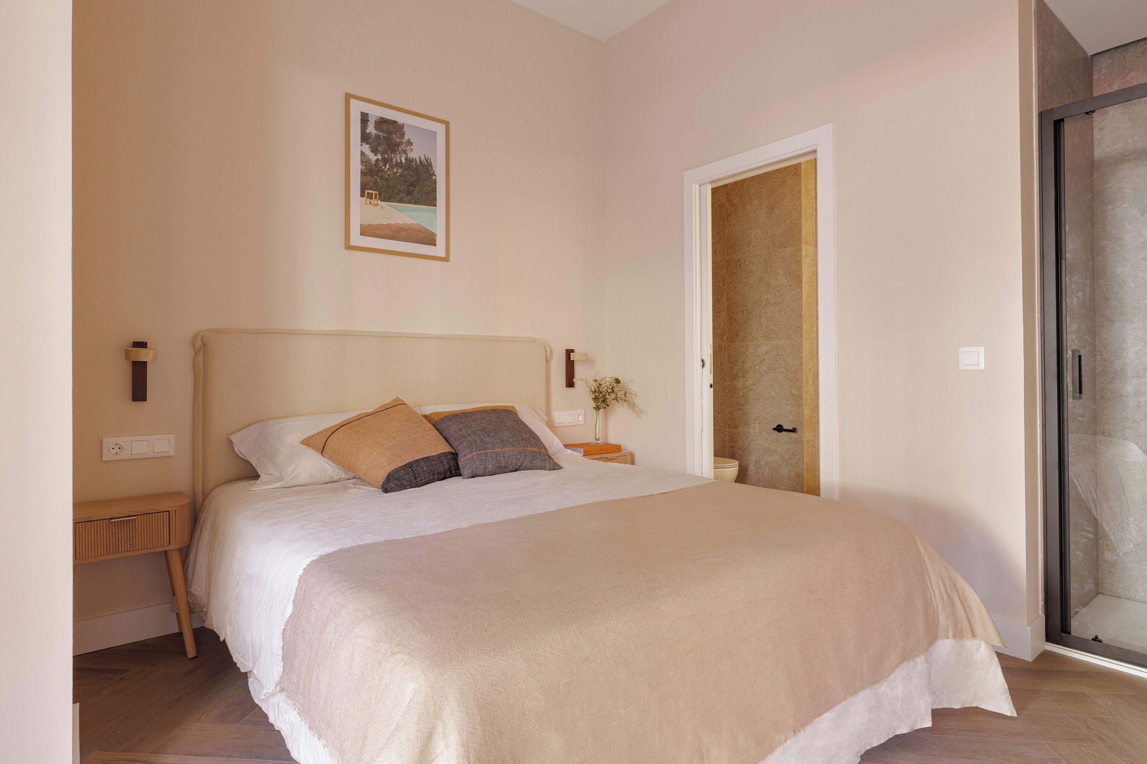 A bedroom with a bed, bedside tables, a nightstand, a framed picture on the wall, and a door leading to another room.