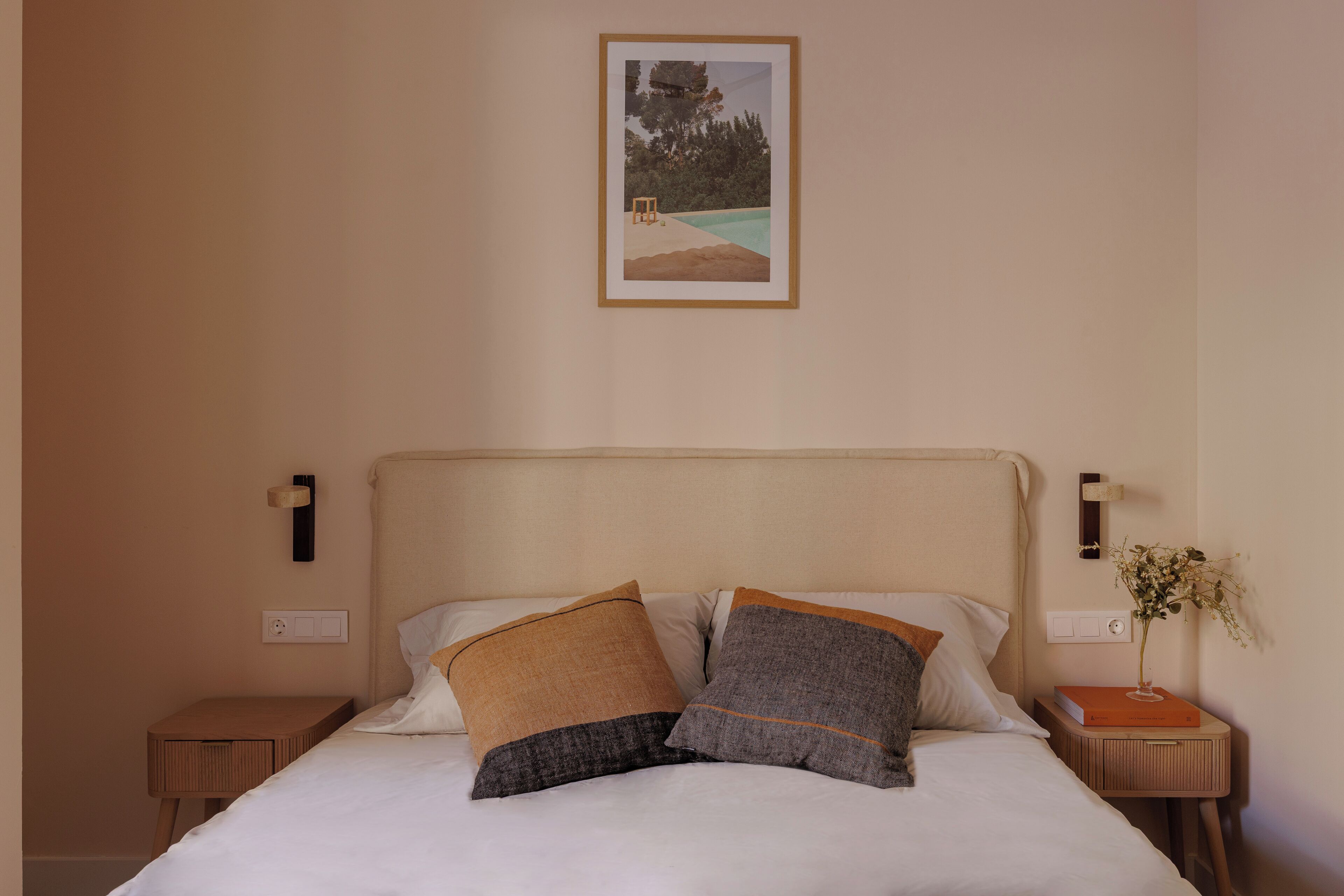 A neatly made bed with two pillows, a bedside table with a book and a vase, and a framed picture on the wall.