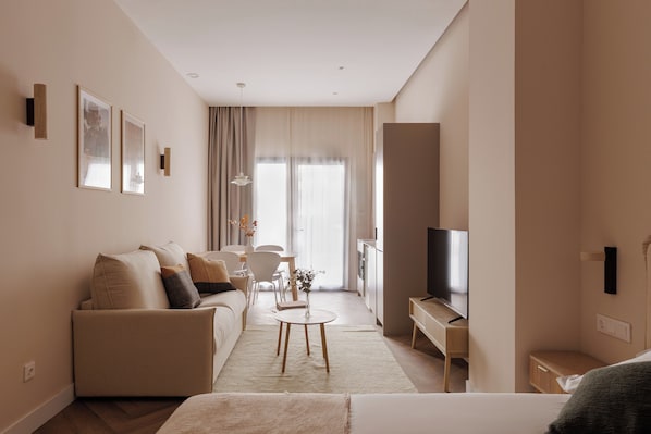 A modern hotel room with a sofa, a small round table, a TV, and a large window.