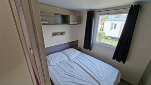 A single bed with a headboard, a bedside table, a window with curtains, and a built-in shelf above the bed. - Mobile home Bungalow Port-la-Nouvelle (Port-la-Nouvelle)