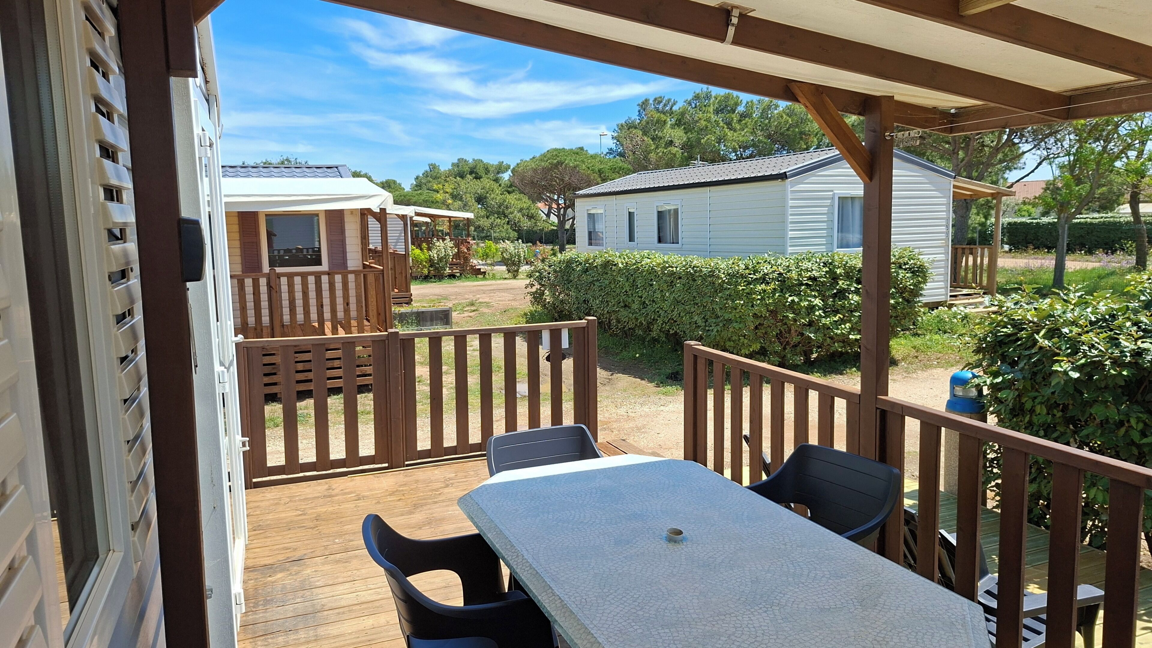 A covered patio with a table and chairs, wooden railings, and a view of a mobile home and garden.