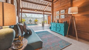 A cozy room with a sofa, a lamp, a blue cabinet, and a view of an outdoor area with plants and a stone wall. - Deluxe Room with Pool Access (Uluwatu)