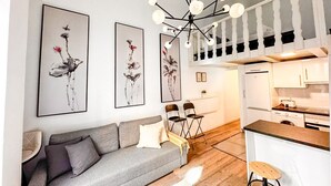 Una sala de estar moderna con un sofá gris, sillas negras, una mesa de comedor de madera y una zona de cocina con armarios blancos y un refrigerador blanco.
