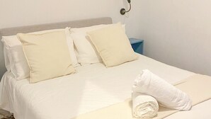 Una cama con ropa blanca, una toalla doblada y una mesita de noche con una lámpara.