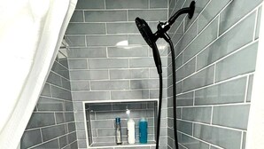 A shower with a black hose, grey tiled walls, and a shelf with toiletries.