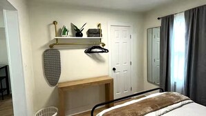 A bedroom with a bed, a bedside table, a hanging laundry rack, a door, and a window with curtains.