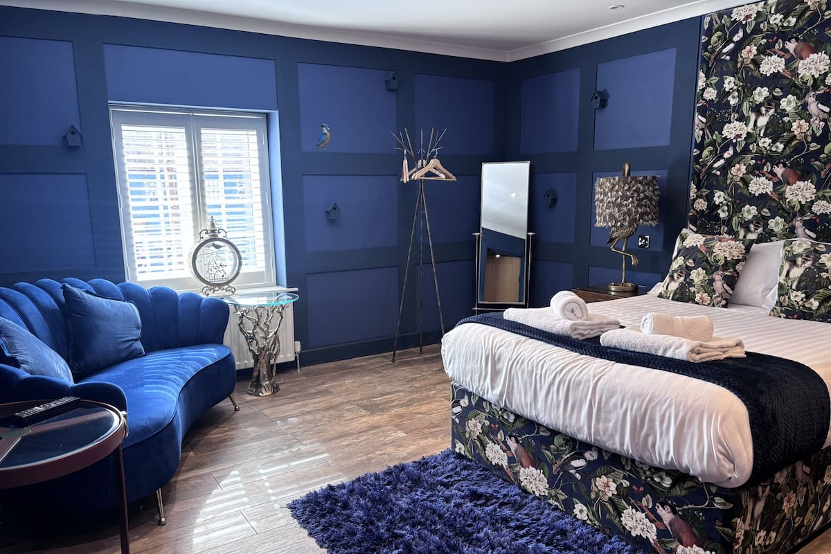 A bedroom with a blue sofa, a bed with floral bedding, a mirror, and a window with blinds.